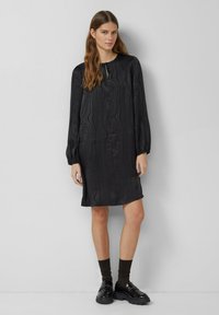 Schwarzes, langärmliges Kleid mit strukturiertem Wellenmuster, rundem Ausschnitt und leicht ausgestellter A-Linien-Form. Kombiniert mit dunklen Socken und glänzenden Schuhen.