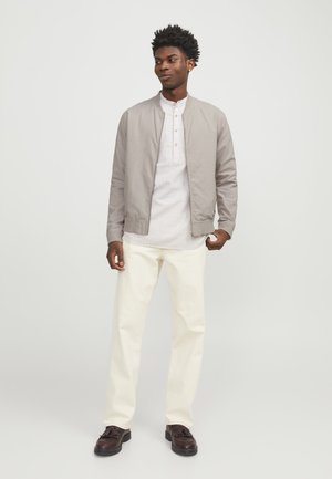 Bomber de color gris claro sobre una camisa blanca con textura, combinado con pantalones crema y zapatos marrón oscuro. Diseño sencillo con un ajuste casual.