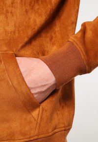 Sudadera de ante marrón con bolsillo frontal, puños acanalados y textura suave. El diseño es casual con un corte holgado.