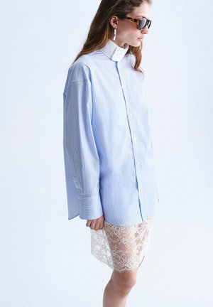 Mujer con camisa de rayas azul claro de gran tamaño sobre una falda de encaje blanca, con gafas de sol de carey y pendientes de aro plateados, vista de perfil.