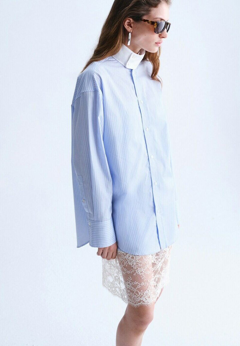 Femme portant une chemise oversize à rayures bleu clair sur une jupe en dentelle blanche, avec des lunettes de soleil écaille de tortue et des boucles d'oreilles argentées en forme d'anneau, vue de profil.