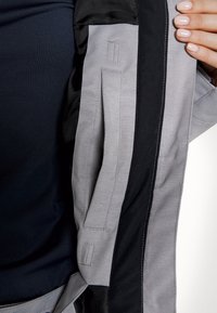 Main tenant ouverte une veste grise et noire côté intérieur, montrant les détails de couture et une poche, avec une chemise bleu foncé en dessous.