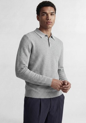 Homme portant un pull polo gris clair à manches longues et un pantalon bleu foncé à fines rayures, debout avec les mains légèrement jointes.