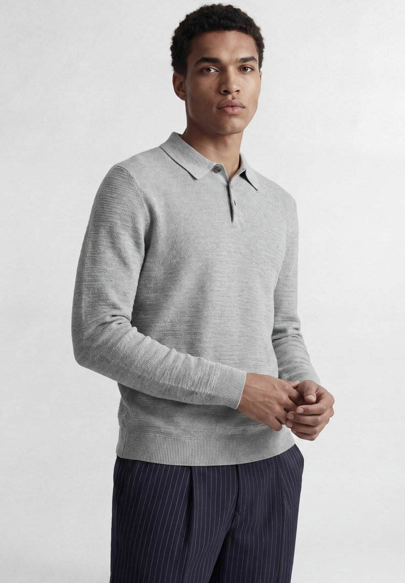 Homme portant un pull polo gris clair à manches longues et un pantalon bleu foncé à fines rayures, debout avec les mains légèrement jointes.