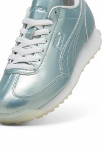 Baskets métalliques bleu clair avec lacets blancs et semelle en caoutchouc. Présente un panneau latéral texturé et un logo embossé sur la languette et le côté.