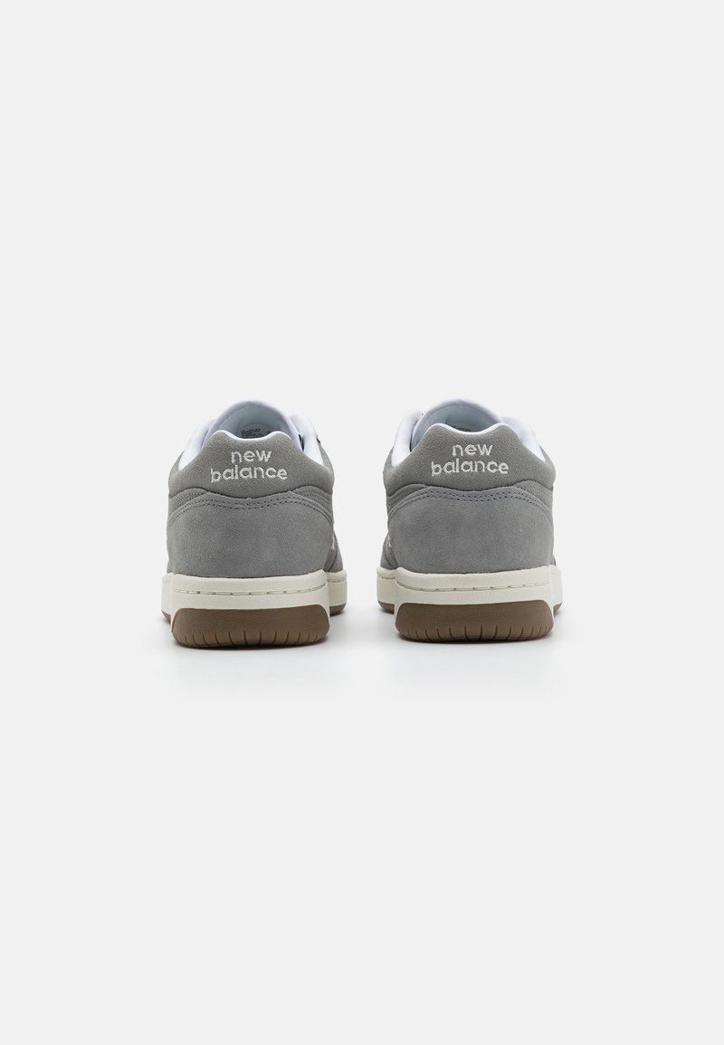 Baskets en suède gris avec des accents blancs, dotées d'un talon rembourré, d'une semelle en caoutchouc et d'un logo "New Balance" en relief à l'arrière.
