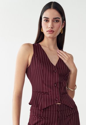 Mujer con cabello largo y oscuro lleva un chaleco sin mangas de rayas finas en color granate y pantalones a juego, complementado con pendientes y pulsera dorados.
