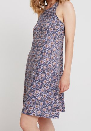 Vrouw draagt een mouwloze, knie-lange blauwe jurk met een roze en witte bloemenprint, staande tegen een effen achtergrond.