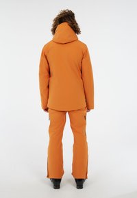 Chaqueta impermeable naranja con capucha y pantalones a juego, que cuentan con bolsillos con cremallera y una textura suave, mostrada desde atrás.