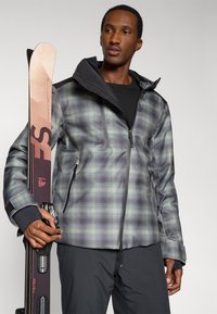 Chaqueta de nieve para hombre con un patrón de cuadros en gris y verde azulado, con cuello alto, bolsillos con cremallera y un diseño elegante y ajustado. Él sostiene esquís.