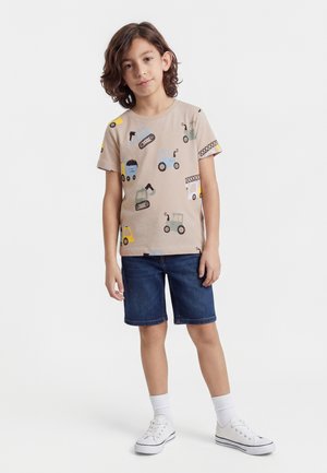 Zēns ar plecu garuma brūniem matiem valkā bēša krāsas T-kreklu ar būvmašīnu apdruku, džinsa šortus, baltas zeķes un baltus sporta apavus.