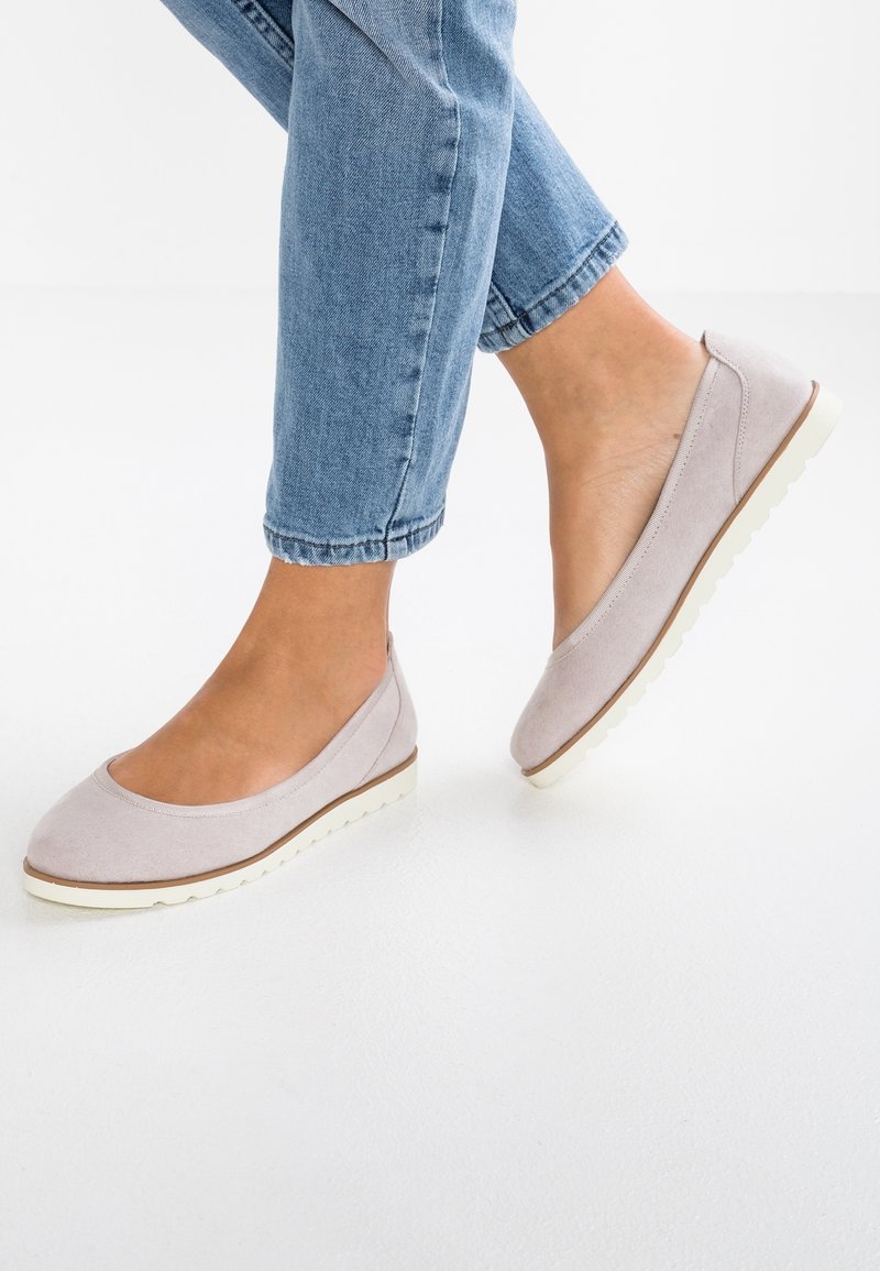 Lys grå ruskindsballerinaer med en rund tå, hvid gummisål og en diskret brun accentlinie langs bunden. Båret med blå jeans.