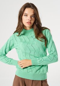 Suéter de punto en verde menta con cuello y puños acanalados, que presenta un patrón ondulado y detallado texturizado en toda la tela.