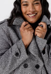 Dubbelzijdige grijze wollen jas met een gestructureerde afwerking, grote kraag en zwarte knopen. Het model houdt de kraag met beide handen vast.
