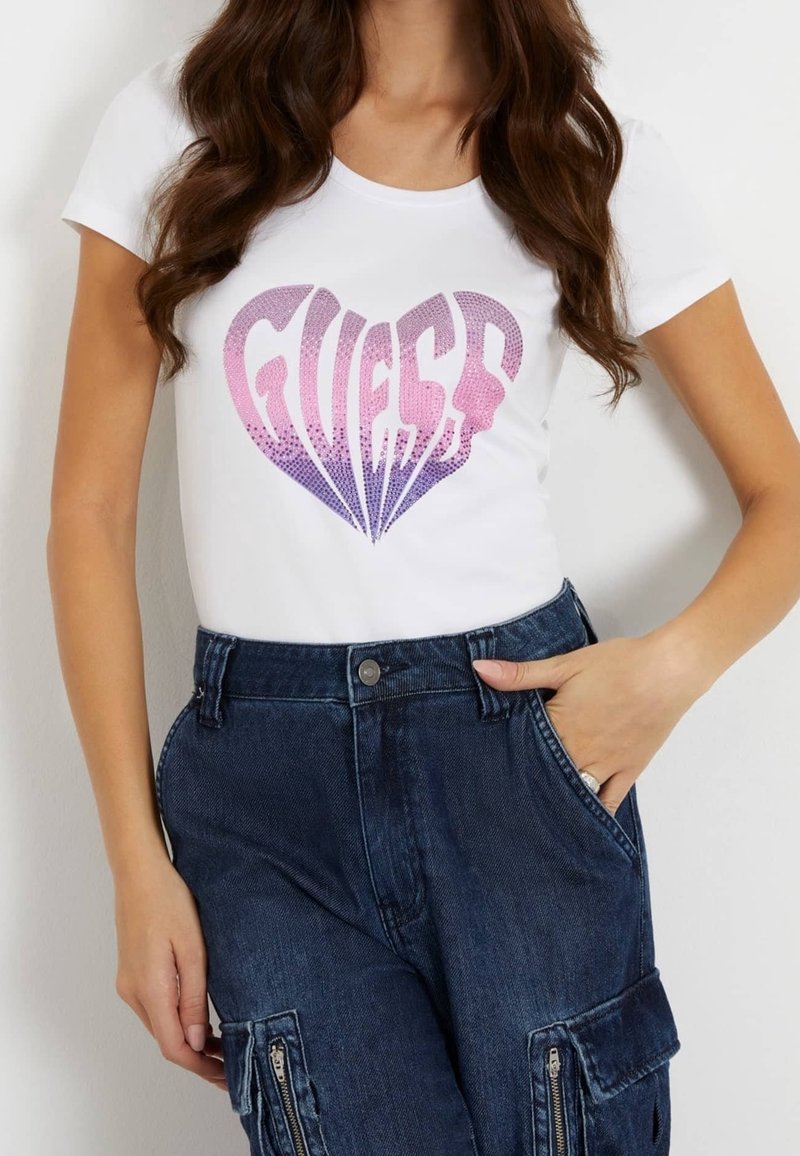 Femme portant un t-shirt blanc à manches courtes avec un logo en forme de cœur rose et violet "GUESS" et un jean cargo bleu foncé taille haute.