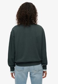 Sudadera verde oscuro con cuello redondo y puños acanalados, combinada con jeans de mezclilla azul claro. Tejido suave; diseño sencillo y relajado.