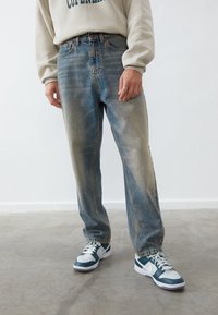 Persona con jeans azules desgastados, sudadera beige y zapatillas blancas y verde oscuro, de pie sobre un suelo de concreto gris.