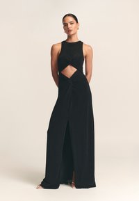 Musta hihaton mekko, jossa on kiertynyt etuosan muotoilu, aukaistu vyötärön kohdalla, pitkä hulmuava helma ja reidestä korkea halkio. Sileä kangaspinta.