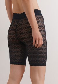 Pantalones cortos de encaje negro con patrón floral en forma de diamante, de longitud hasta la mitad del muslo, cintura con borde en forma de ondas y textura suave. Presentan un diseño ajustado.