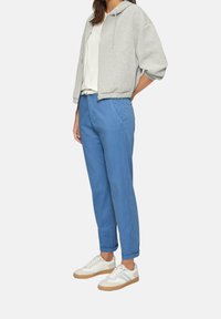 Szara krótka bluza z kapturem, biała koszulka, niebieskie zwężane spodnie i białe trampki z beżowymi akcentami. Luźny strój na modelce.