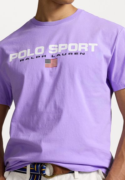 Camiseta de algodón lila con "POLO SPORT" y "RALPH LAUREN" en letras blancas en negrita, con un pequeño gráfico de la bandera americana debajo.