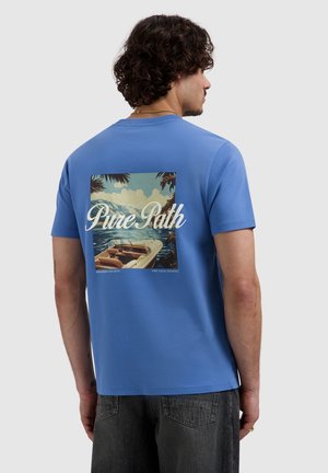 Man met krullend haar die een blauw t-shirt draagt met een rugprint van een boot op het water met bergen en de tekst "Pure Path."