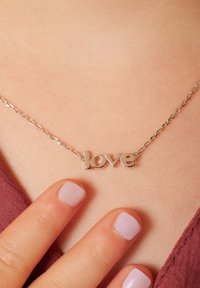Zilveren ketting met een "liefde" hanger. De hanger heeft een gladde, reflecterende oppervlakte. Nagels zijn licht lila geverfd.