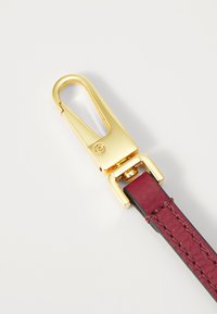 Guldfarvet metalclips fastgjort til et bordeaux læderrem med en tekstureret overflade, der har syninger langs kanterne.