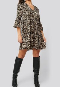 Kleid mit Leopardenmuster, V-Ausschnitt, dreiviertellangen Rüschem Ärmeln, einem gestuften Design und einer fließenden Silhouette. Kombiniert mit knielangen schwarzen Stiefeln.