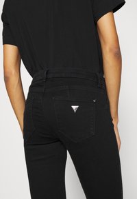 Jeans de mezclilla negra con cintura alta, que cuentan con bolsillos traseros y un detalle de logo metálico triangular. Textura suave y diseño ajustado.