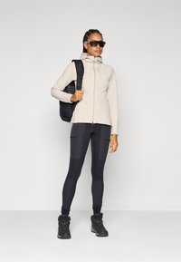 Veste en polaire beige zippée, leggings noirs avec poches et bottes de randonnée noires. Porte un sac à dos noir et des lunettes de soleil foncées.