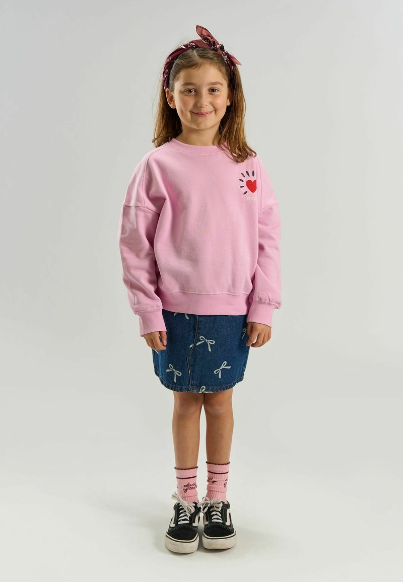 Jeune fille souriante, portant un sweat-shirt rose avec un motif en forme de cœur, une jupe en jean avec des nœuds, des chaussettes roses, des baskets noires et un bandeau rouge.