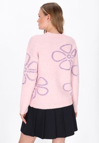 Kobieta ubrana w różowy sweter z purpurowymi wzorami kwiatowymi i detalami z koralików, w połączeniu z czarną plisowaną spódnicą, stojąca bokiem.