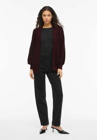 Tumši sarkans adīts kardigans ar gariem piedurknēm, atvērtu priekšpusi un teksturētu rakstu, kombinēts ar melnām biksēm ar augstu jostasvietu un melnām apaviem ar papēdi.