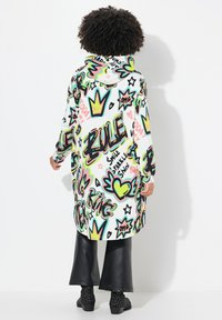 Veste à capuche surdimensionnée avec un imprimé graffiti vibrant et coloré sur fond blanc. Dotée de manches longues et d'une coupe décontractée, accompagnée de pantalons noirs évasés.