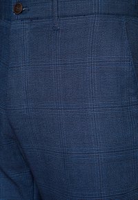 Niebieska tkanina w kratę z bliska, z teksturowanym splocie, przedstawiającym wzór przecinających się linii oraz subtelne różnice w odcieniach. Widoczny detal z guzikiem.