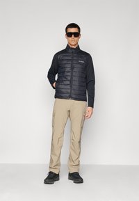 Man wearing black padded jacket, beige cargo pants, black hiking shoes, and large black sunglasses standing against plain background.