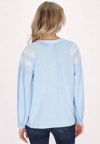 Hellblaue Bluse mit langen Ärmeln und bestickten Akzenten auf den Schultern. Aus strukturiertem Stoff mit lockerer Passform und Knopfmanschetten.