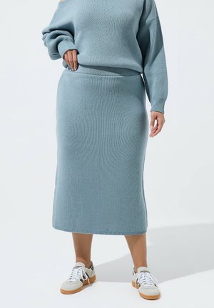 Femme portant un pull en maille bleu clair et une jupe midi assortie, associée à des baskets blanches, debout devant un fond uni clair.