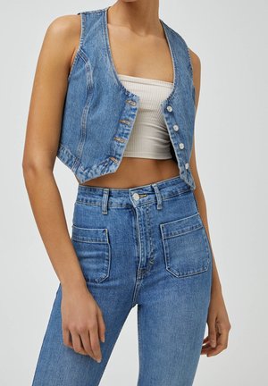 Frau trägt ärmellose blaue Jeansweste über weißem geripptem bauchfreiem Top und hochtaillierter blauer Jeans mit vorderen aufgesetzten Taschen.
