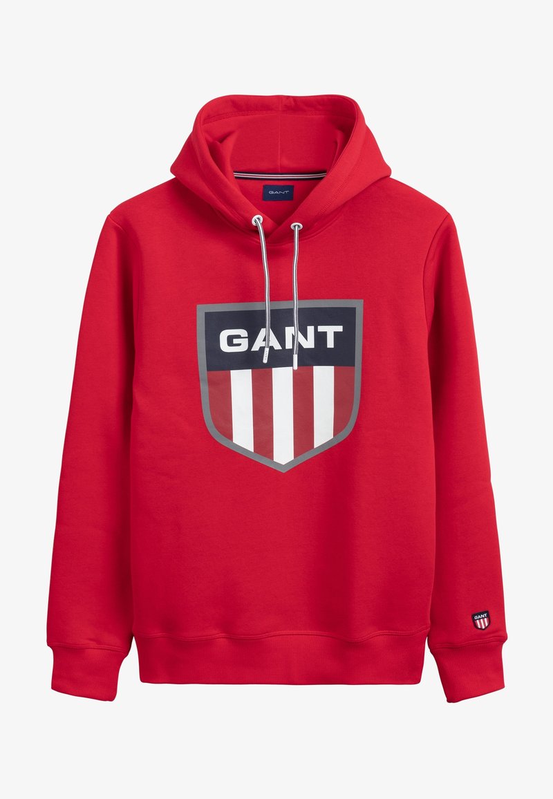 Sweat à capuche rouge avec logo en forme de blason à l'avant, affichant le texte "GANT" au-dessus de rayures verticales rouges et blanches et des cordons sur la capuche.
