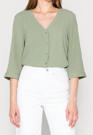 Woman wearing a light green V-neck buttoned blouse with three-quarter sleeves paired with high-waisted white pants.