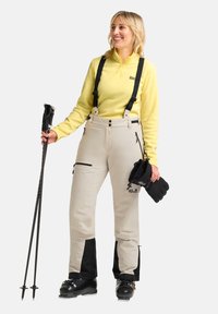 Beige skibroek met zwarte accenten, verstelbare banden, meerdere ritszakken. Gele lange mouwen top. Skistokken en handschoenen in de handen.