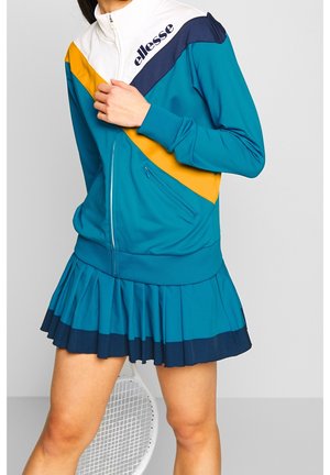 Mujer con chaqueta de chándal azul, amarilla y blanca y falda azul plisada, sosteniendo la cremallera de la chaqueta, raqueta de tenis detrás de sus piernas.