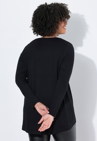 Zwarte lange mouwen top met een ronde halslijn, ontspannen pasvorm, zijsplitten en zachte textuur. Gecombineerd met zwarte aansluitende leggings.