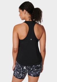Vrouw met krullend haar in een paardenstaart, gekleed in een zwarte racerback tanktop en shorts met een zwart, wit en grijs patroon, die van zich af kijkt.