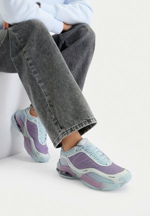 Osoba siedząca w szarych dżinsach i jasnoniebiesko-fioletowych tenisówkach na jednolitym białym tle.