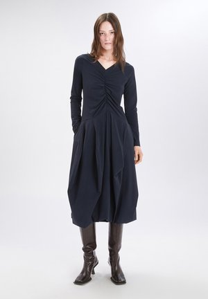 Abito lungo blu navy con maniche lunghe, scollatura a V, parte anteriore arricciata e gonna a pieghe ampia. Il modello indossa stivali marroni al ginocchio. Tessuto di texture liscia.