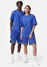 T-shirts en coton bleu avec un logo, associés à des shorts bleus assortis, présentant des accents noirs et blancs. Les deux mannequins portent des baskets blanches.