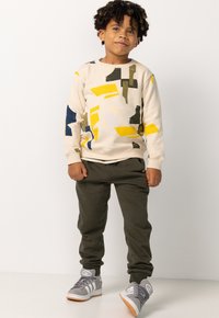 Beige sweatshirt met abstracte gele, groene en blauwe patronen; gecombineerd met olijfgroene sweatpants en grijze sneakers met witte strepen.